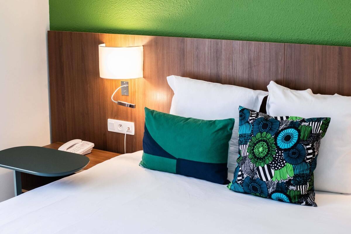 A hotel room with a wooden headboard, a bedside lamp, a telephone, and a bed with green and patterned pillows.
