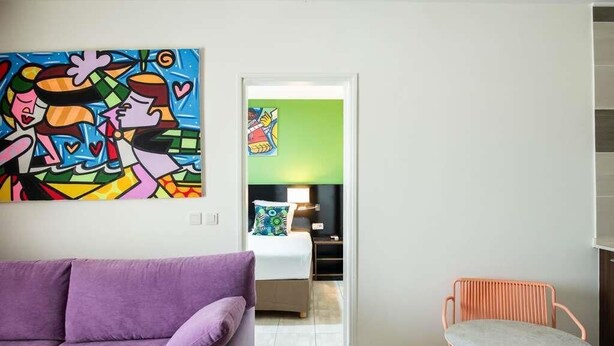 A modern living room with a purple sofa, a round table with orange legs, and a colorful abstract painting on the wall. - Ibis Styles Cayenne Centre Amazonia (Ouverture Juin 2024) (Cayenne)