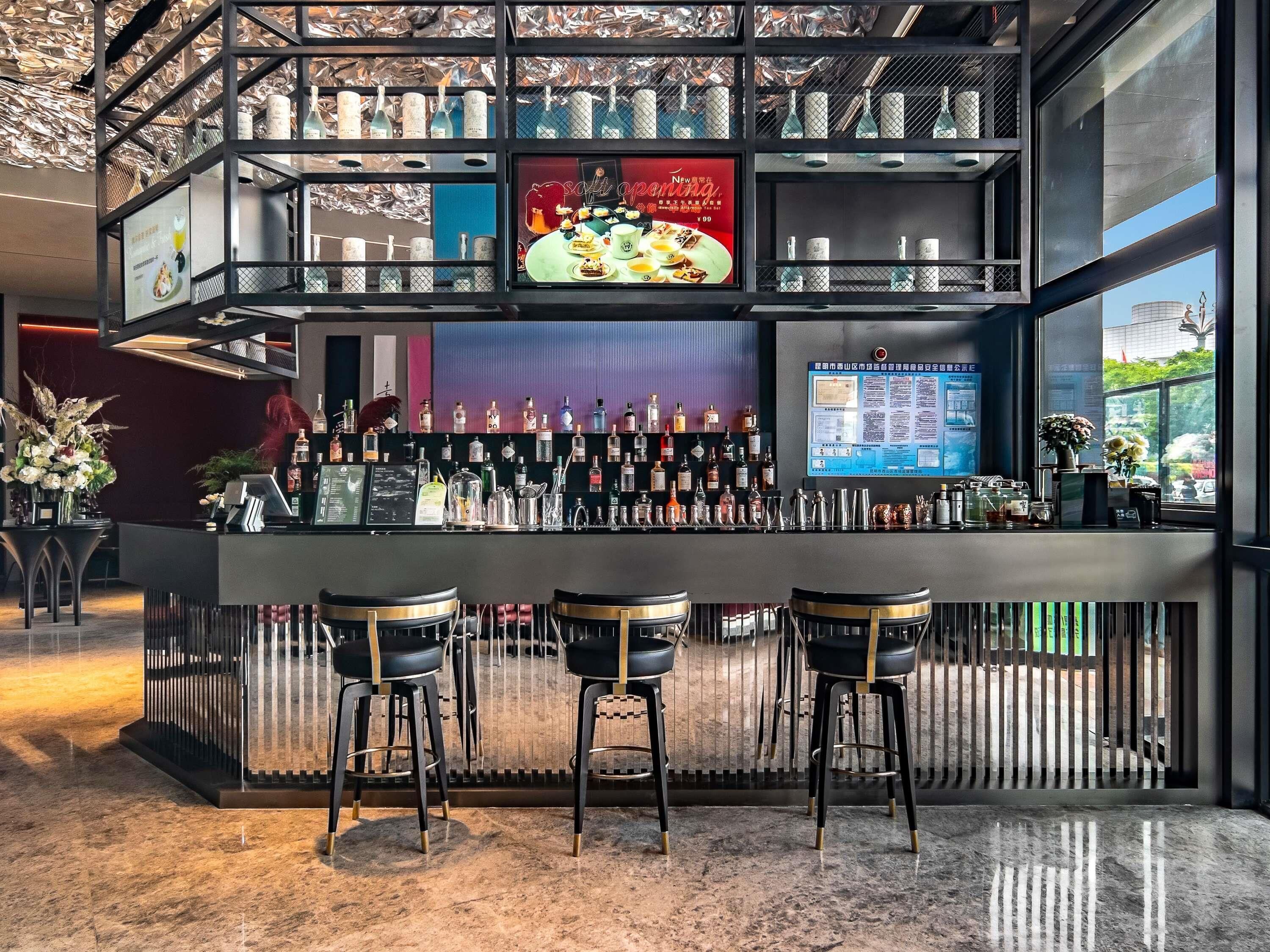 A bar area with a modern design, featuring a counter with multiple bottles and glasses, three stools, and a digital menu display.