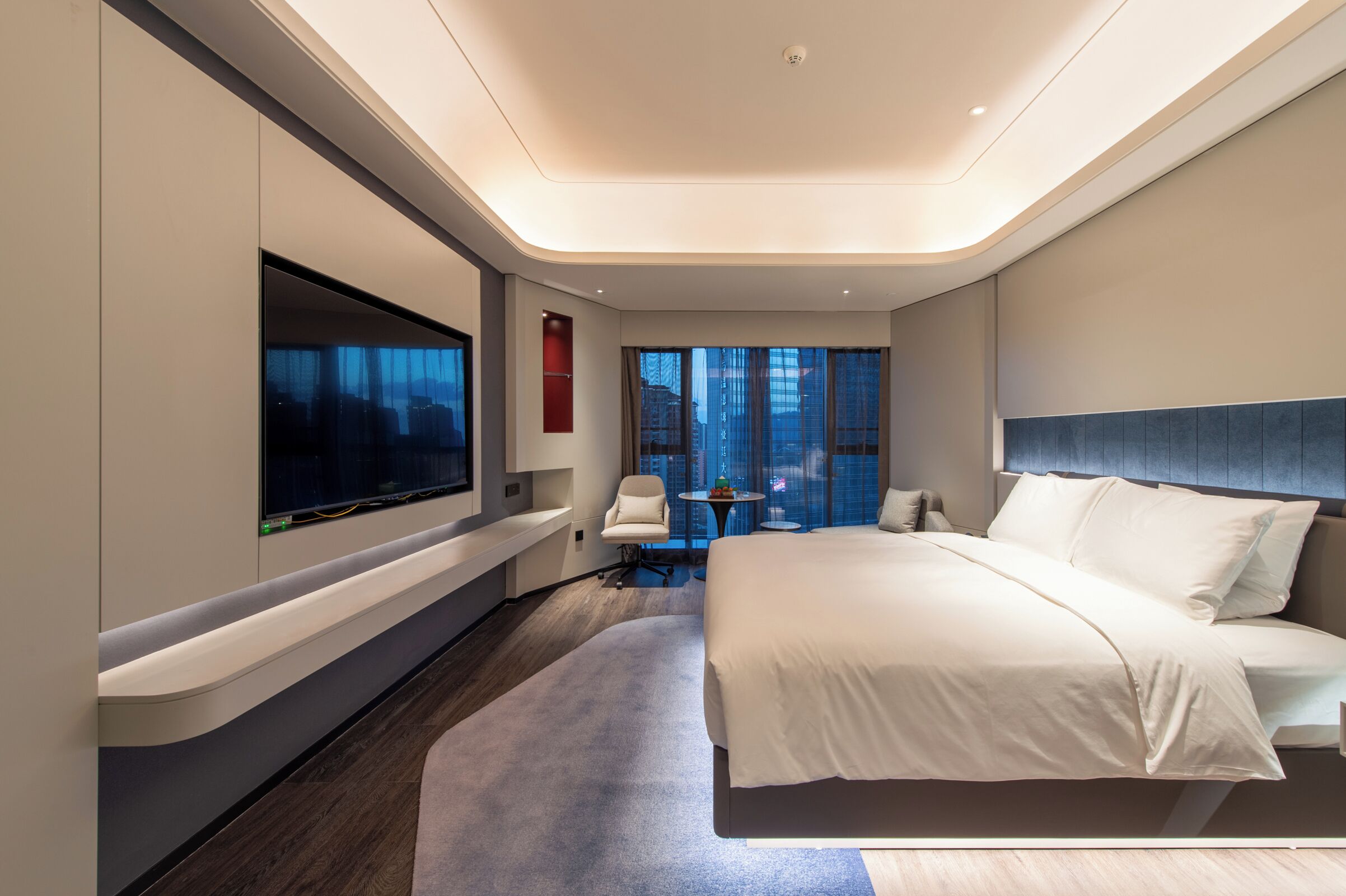 Una habitación de hotel moderna con una cama grande, un televisor de pantalla plana en la pared, una zona de estar con una silla y una mesita, y vistas a la ciudad a través de amplios ventanales.