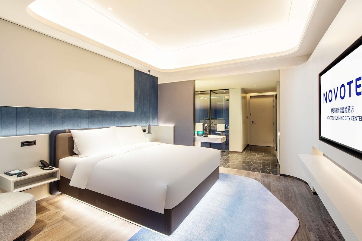 Una habitación de hotel moderna con una cama grande, mesitas de noche, un escritorio y un baño visible a través de un panel de vidrio.