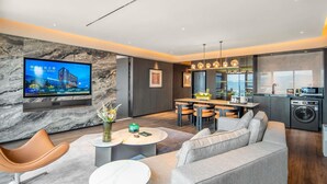 Uma sala moderna com um sofá, uma TV fixada numa parede de mármore, um espaço de refeições com mesa e cadeiras, e uma cozinha com máquina de lavar roupa.