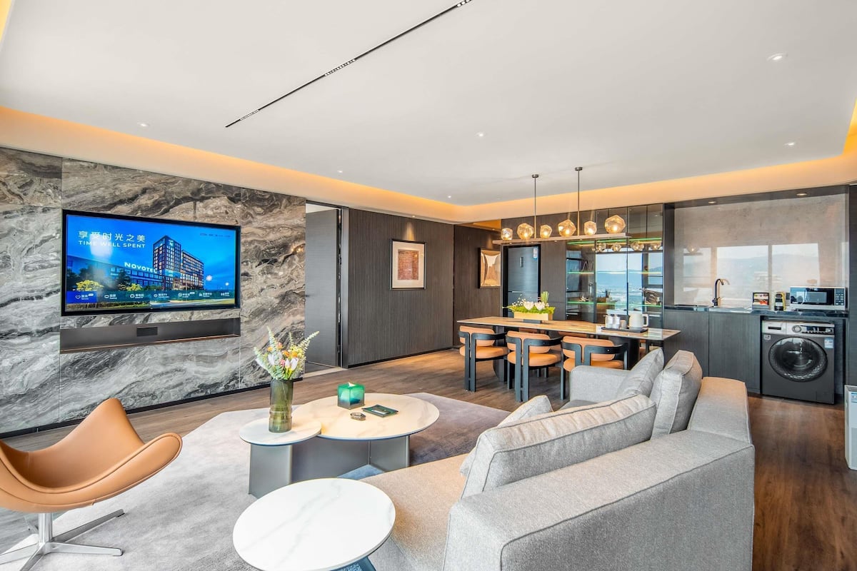 Una sala moderna con sofá, televisor montado en una pared de mármol, comedor con mesa y sillas, y cocina con lavadora.