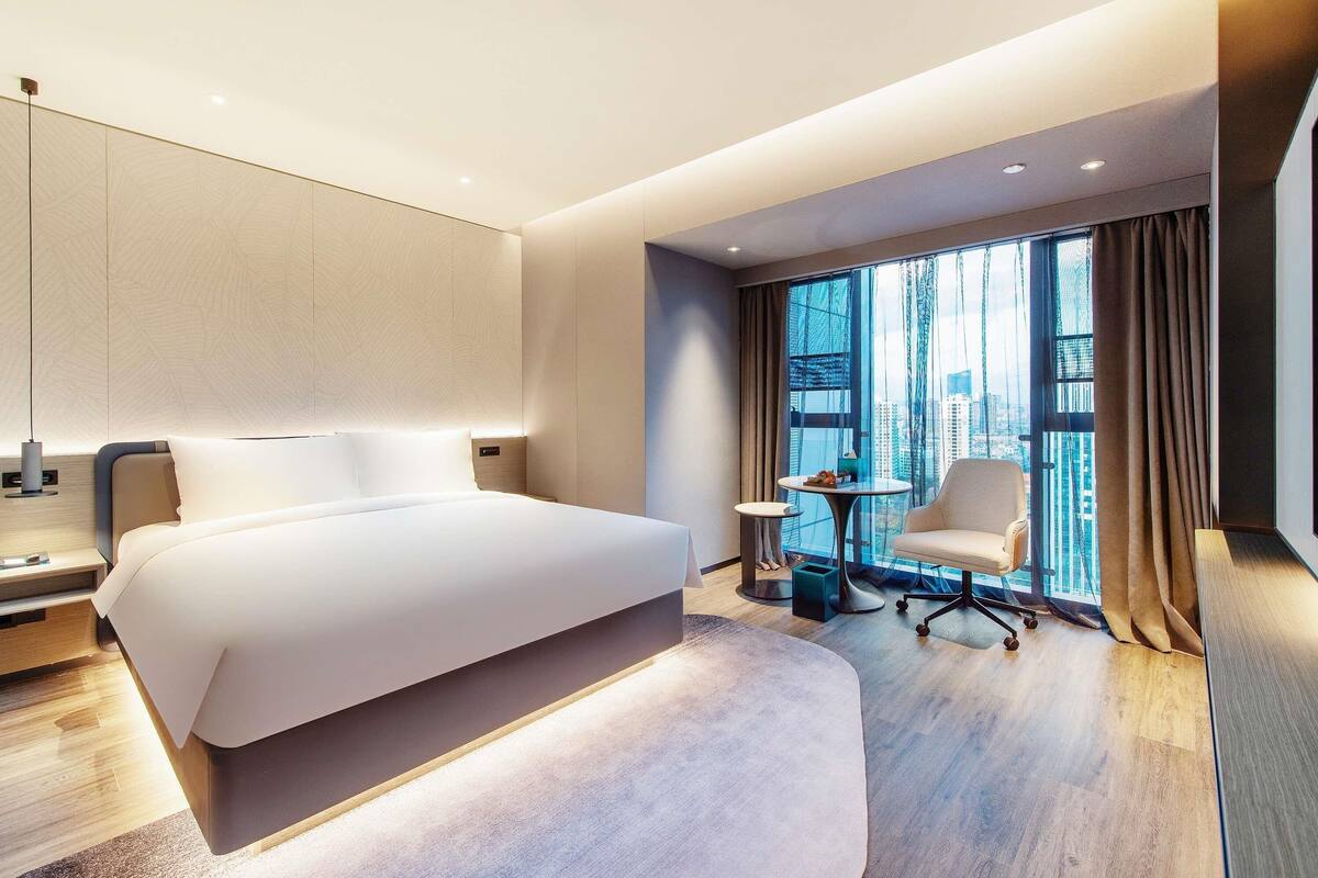 Habitación de hotel moderna con una cama grande, una mesita redonda pequeña, una silla y vistas a la ciudad a través de amplios ventanales.