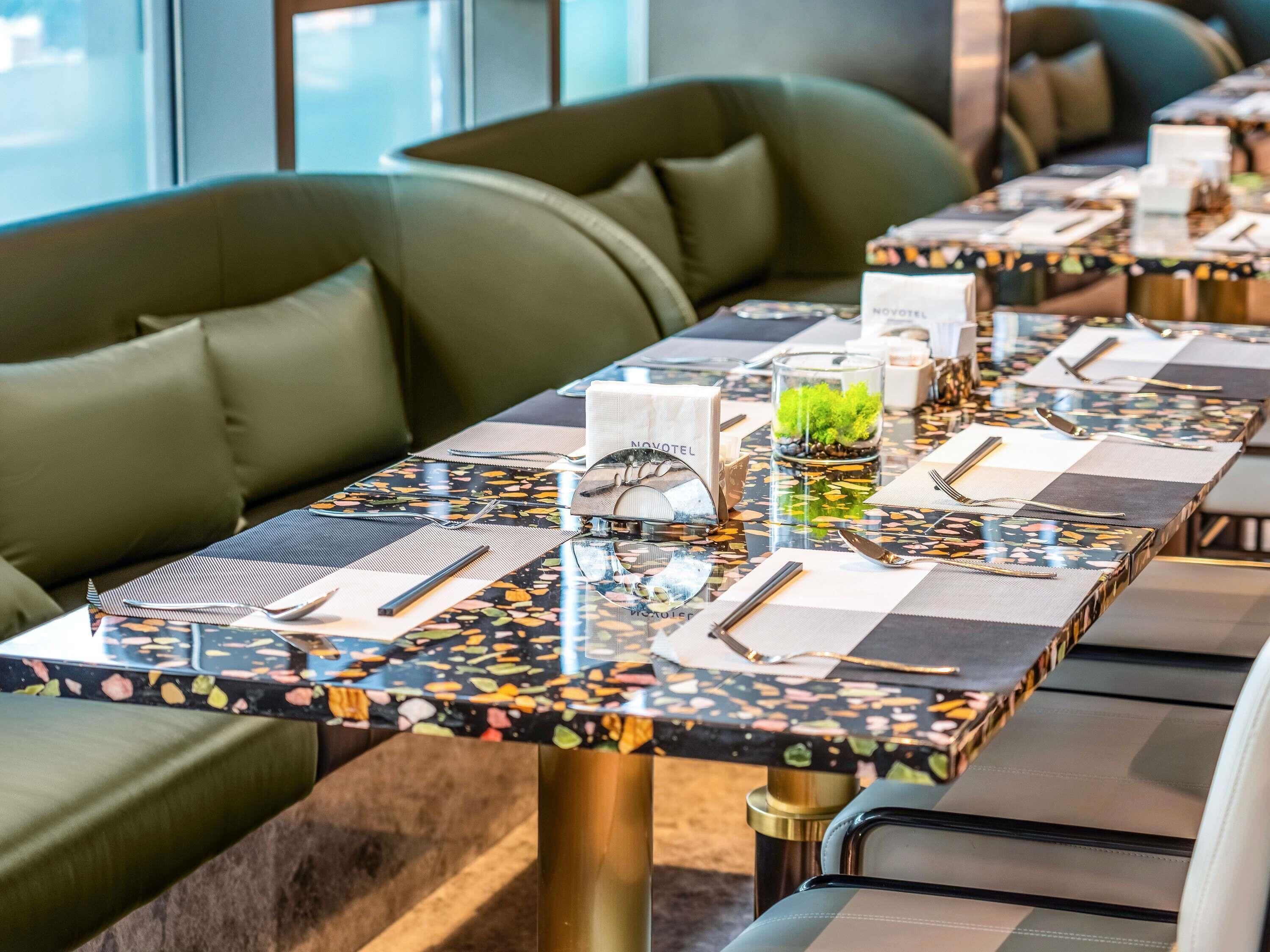 Restaurante com uma mesa comprida posta, incluindo talheres, guardanapos e um recipiente de vidro com uma planta verde.