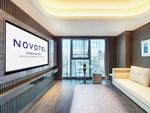 A modern hotel room with a large TV screen displaying 'NOVOTEL KUNMING CITY CENTER,' a city view through large windows, a sofa, and a small round table. - Novotel Kunming City Center (Kunming)