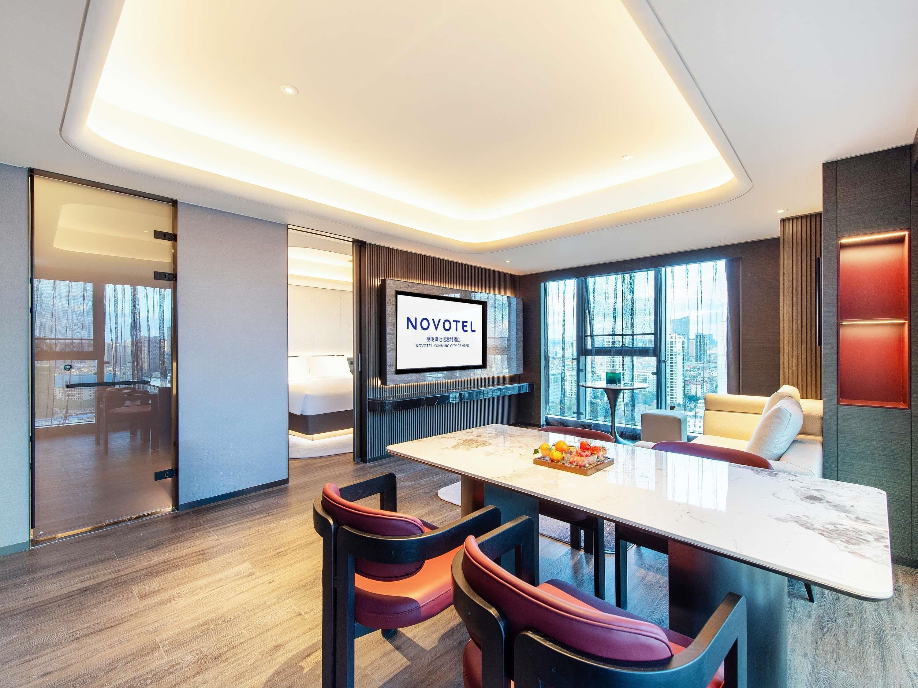 A modern hotel room with a large TV, a dining area with a table and chairs, and a view of the city.