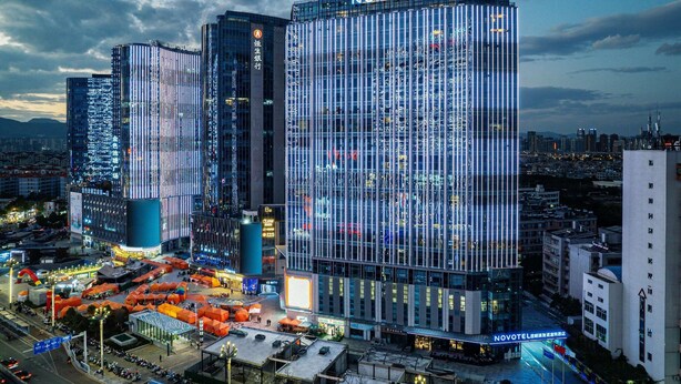 Um hotel moderno com uma placa de 'NOVOTEL' bem visível, situado numa área urbana movimentada, com ruas e outros edifícios.