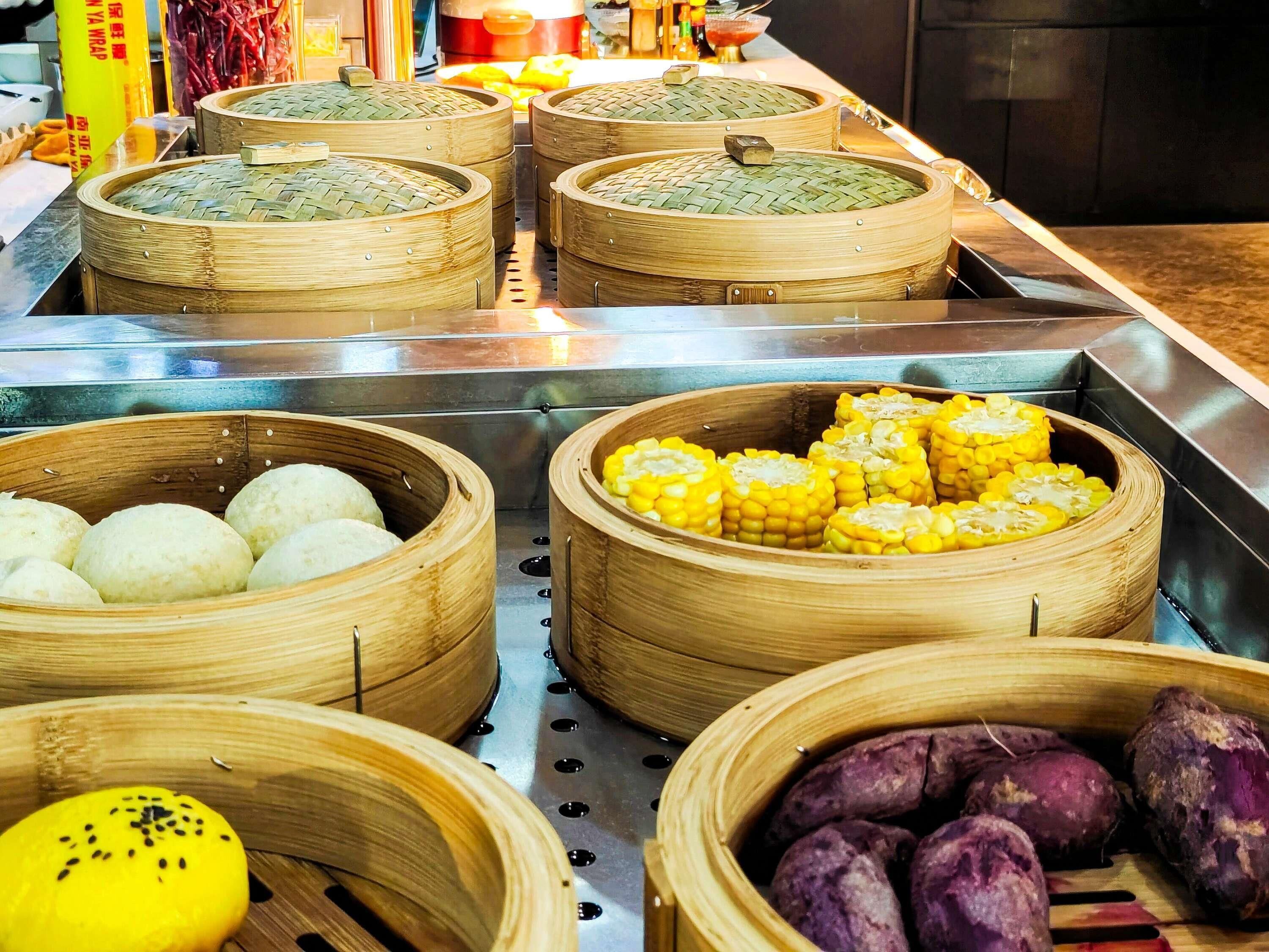 Diversos alimentos en vaporeras de bambú, como dumplings, mazorcas de maíz y batatas moradas.