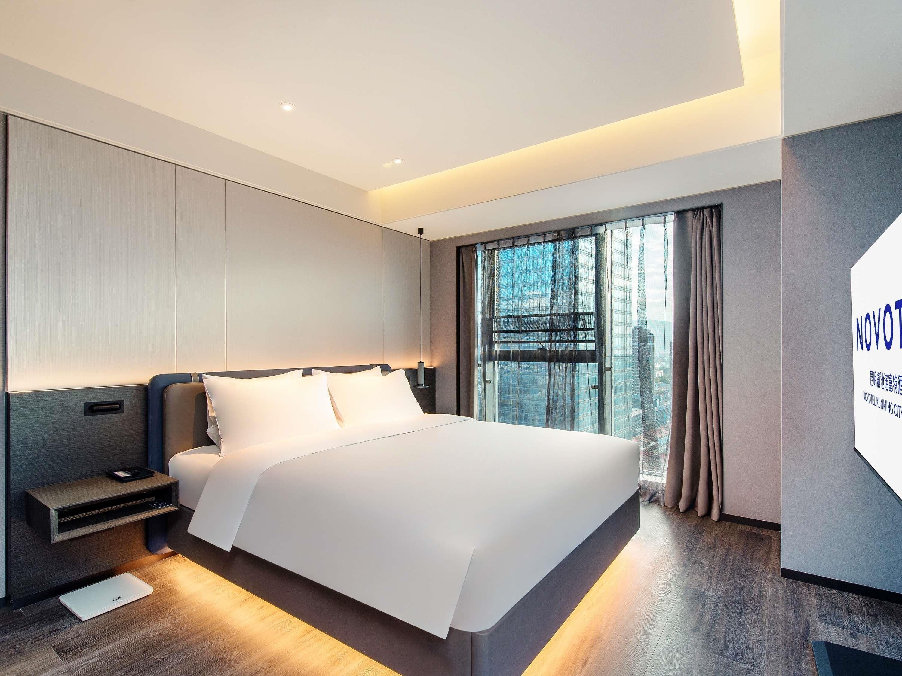 A modern hotel room with a large bed, a bedside table with a lamp, a laptop on the floor, and a city view through the window.