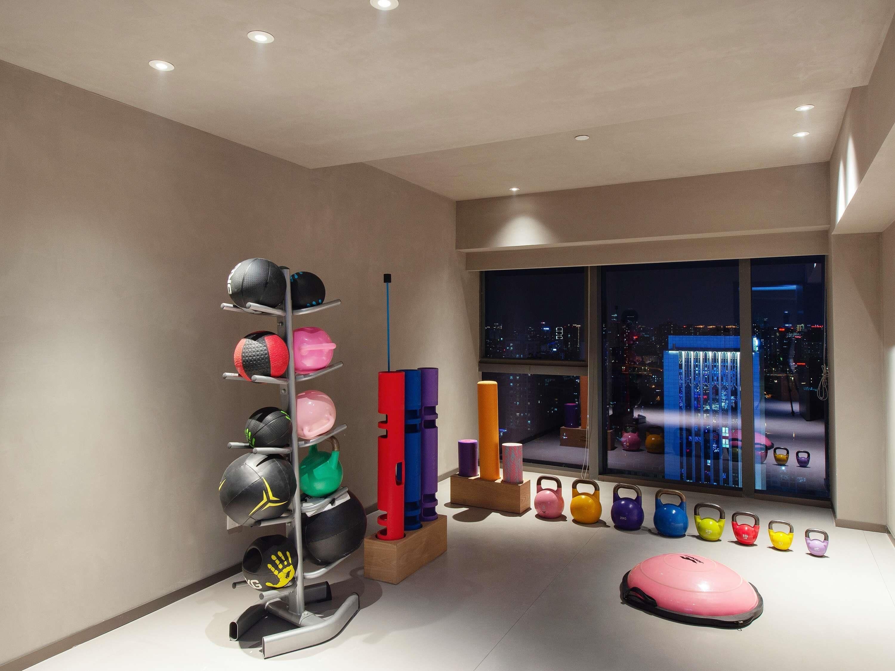 Una habitación moderna con un ventanal grande, un techo con iluminación empotrada y un estante montado en la pared con equipo de ejercicio, además de una colchoneta de ejercicio rosa.