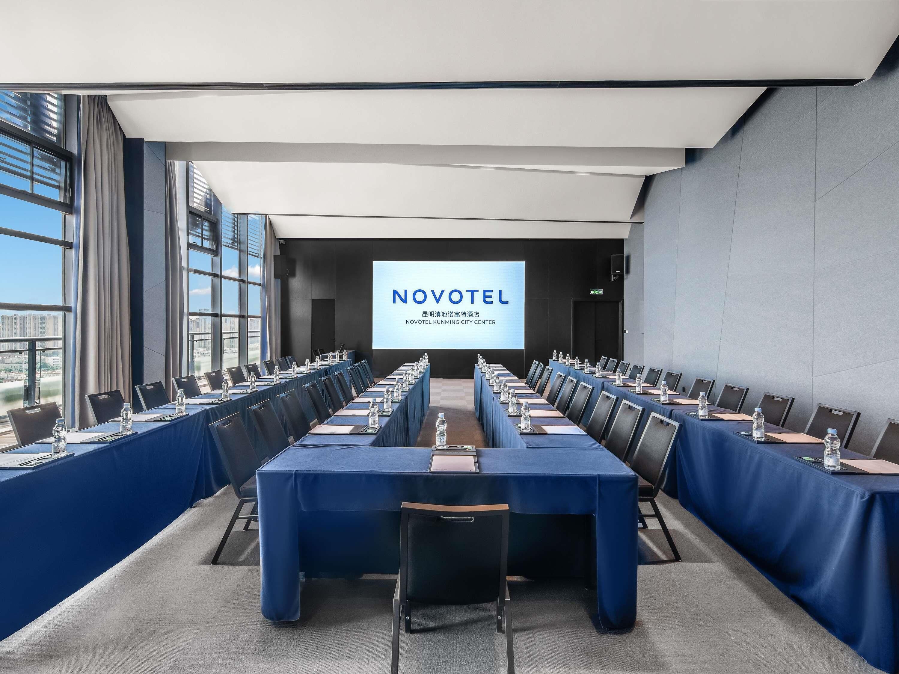A conference room with long tables, chairs, and a projection screen displaying 'NOVOTEL'.