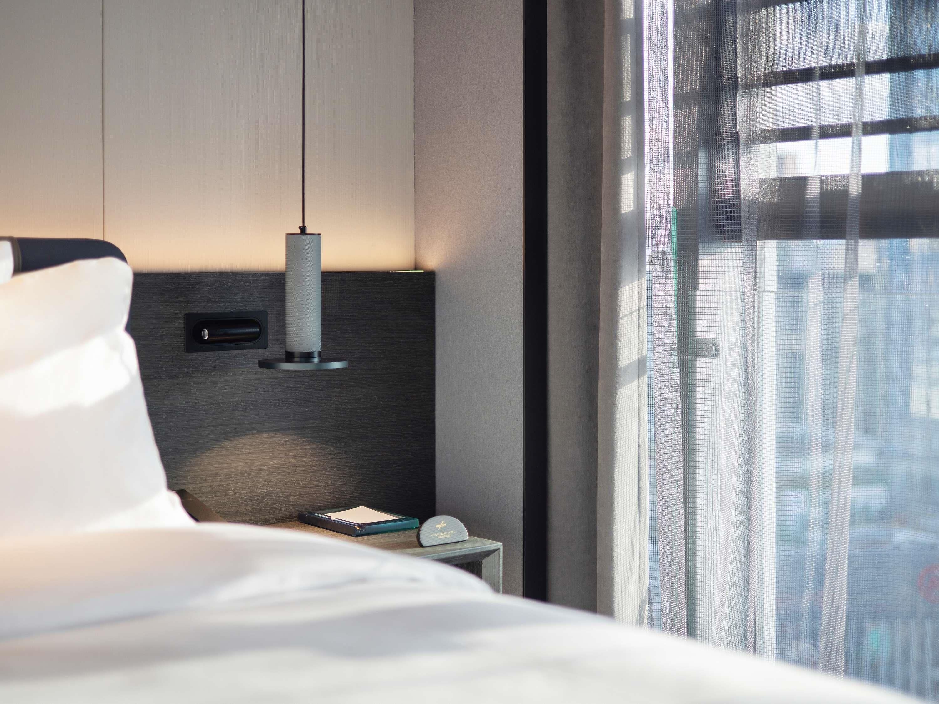 A modern hotel room with a bed, a bedside table with a lamp, a small clock, and a window with a view of buildings.