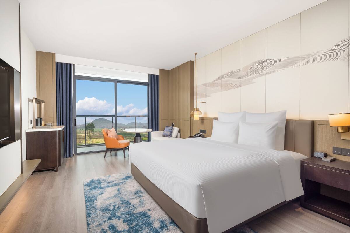 Ein modernes Hotelzimmer mit einem großen Bett, einem Schreibtisch, einem Sessel und Blick ins Freie.