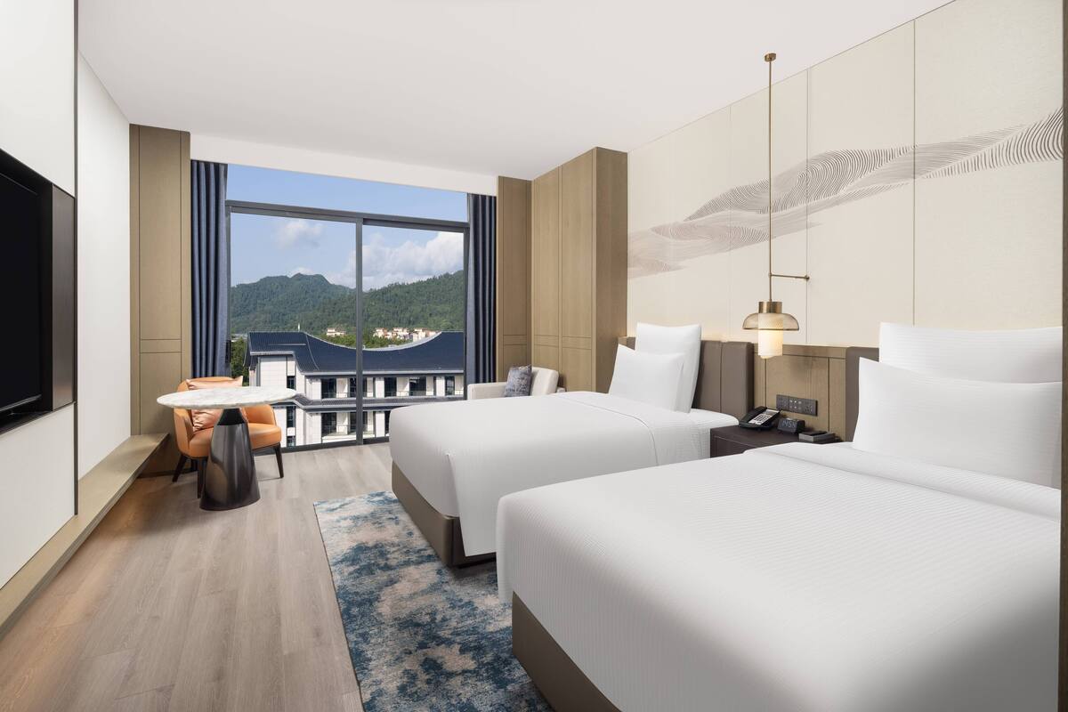 Ein Hotelzimmer mit zwei Betten, einem Schreibtisch, einem Stuhl, einem Fernseher und einem großen Fenster mit Blick auf Berge und Gebäude.
