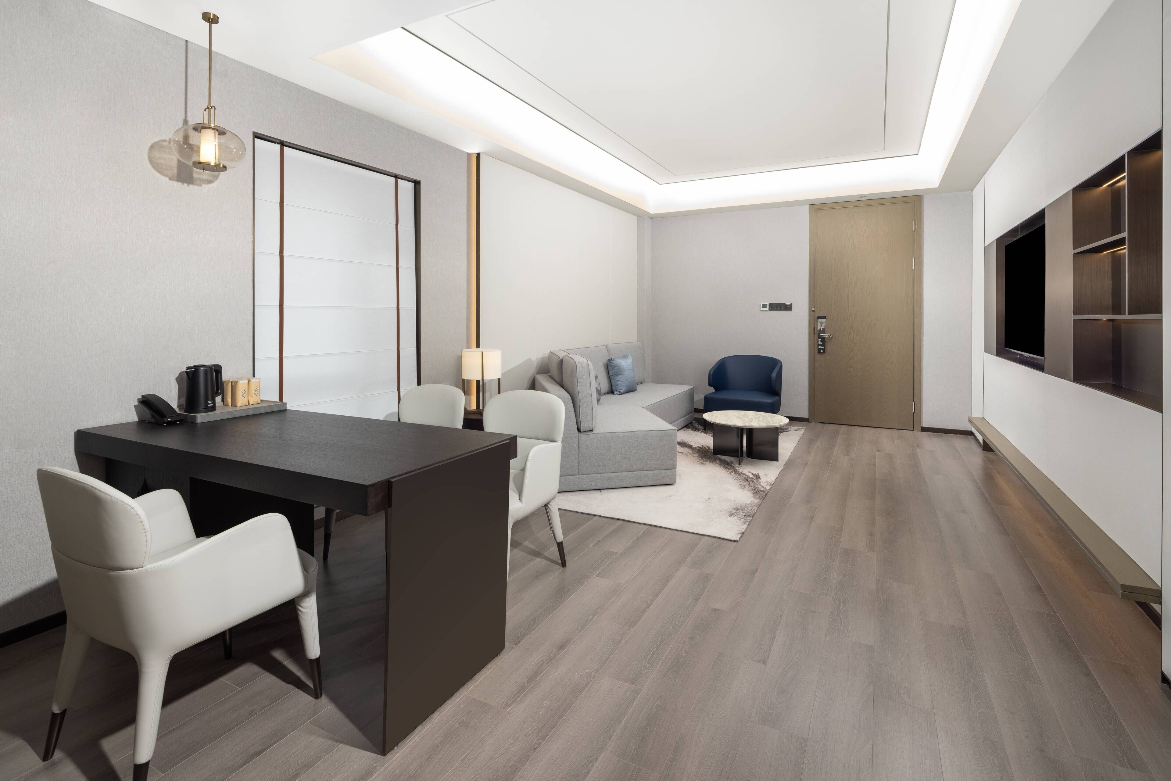 A modern hotel room with a dark wooden desk, white chairs, a grey sofa, and a small round table.