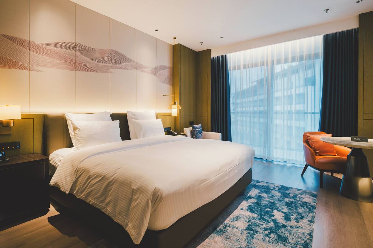 Ein modernes Hotelzimmer mit einem großen Bett, einem Schreibtisch, einem Sessel und Blick auf die Stadt durch große Fenster.