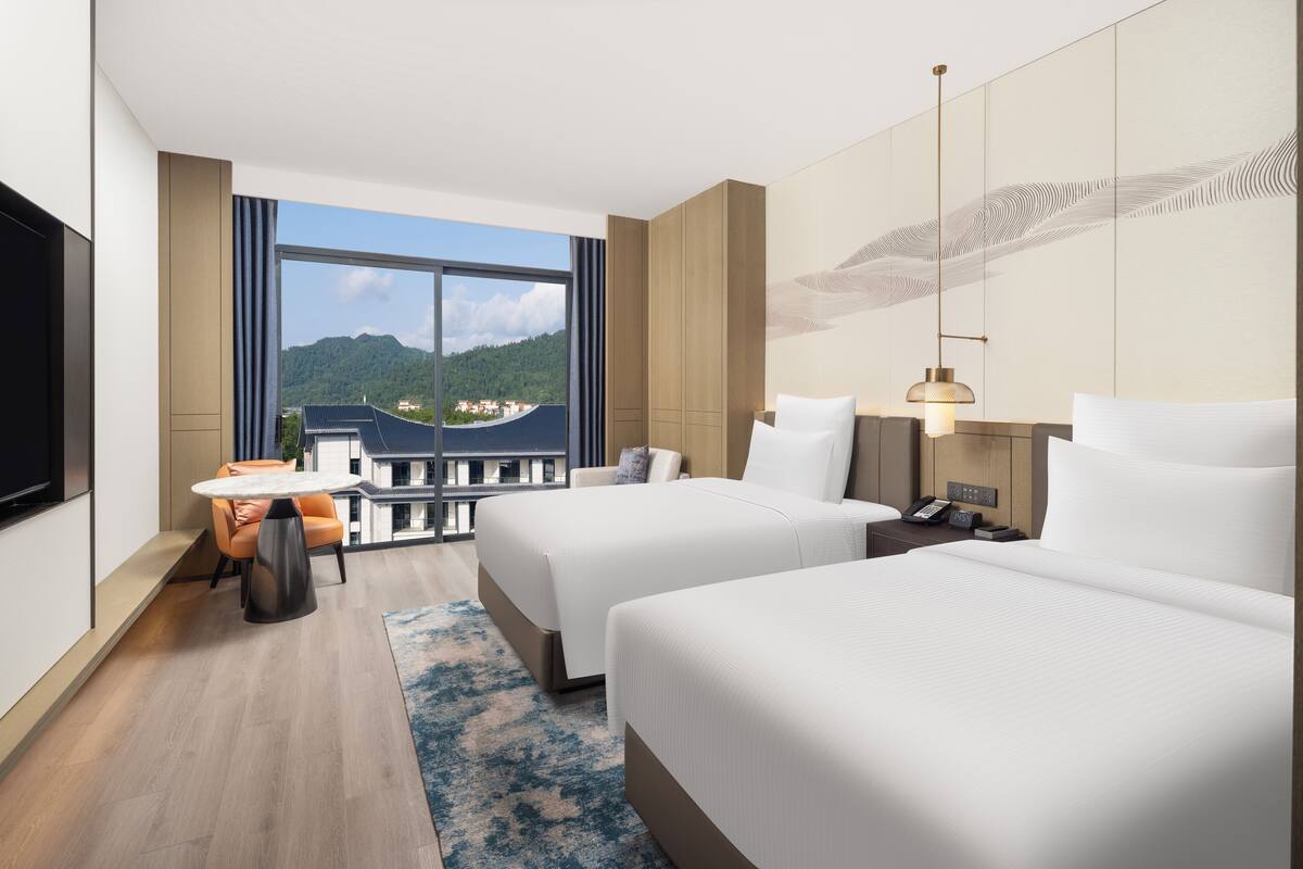 Ein Hotelzimmer mit zwei Betten, einem Schreibtisch, einem Sessel, einem Fernseher und einem großen Fenster mit Blick auf einen Berg und Gebäude.
