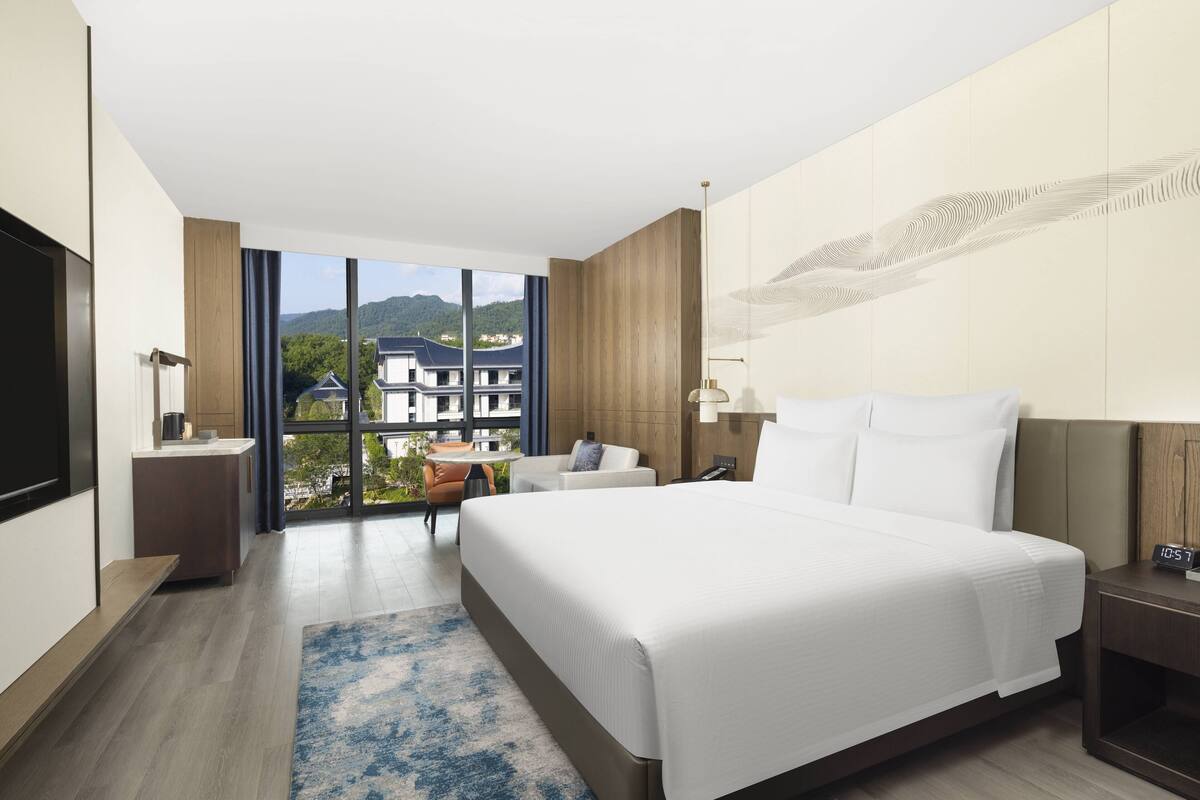 Ein modernes Hotelzimmer mit einem großen Bett, einem Schreibtisch, einem Sessel, einem Fernseher und Blick auf Gebäude und Berge.