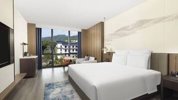 A modern hotel room with a large bed, a desk, a chair, a TV, and a view of buildings and mountains.