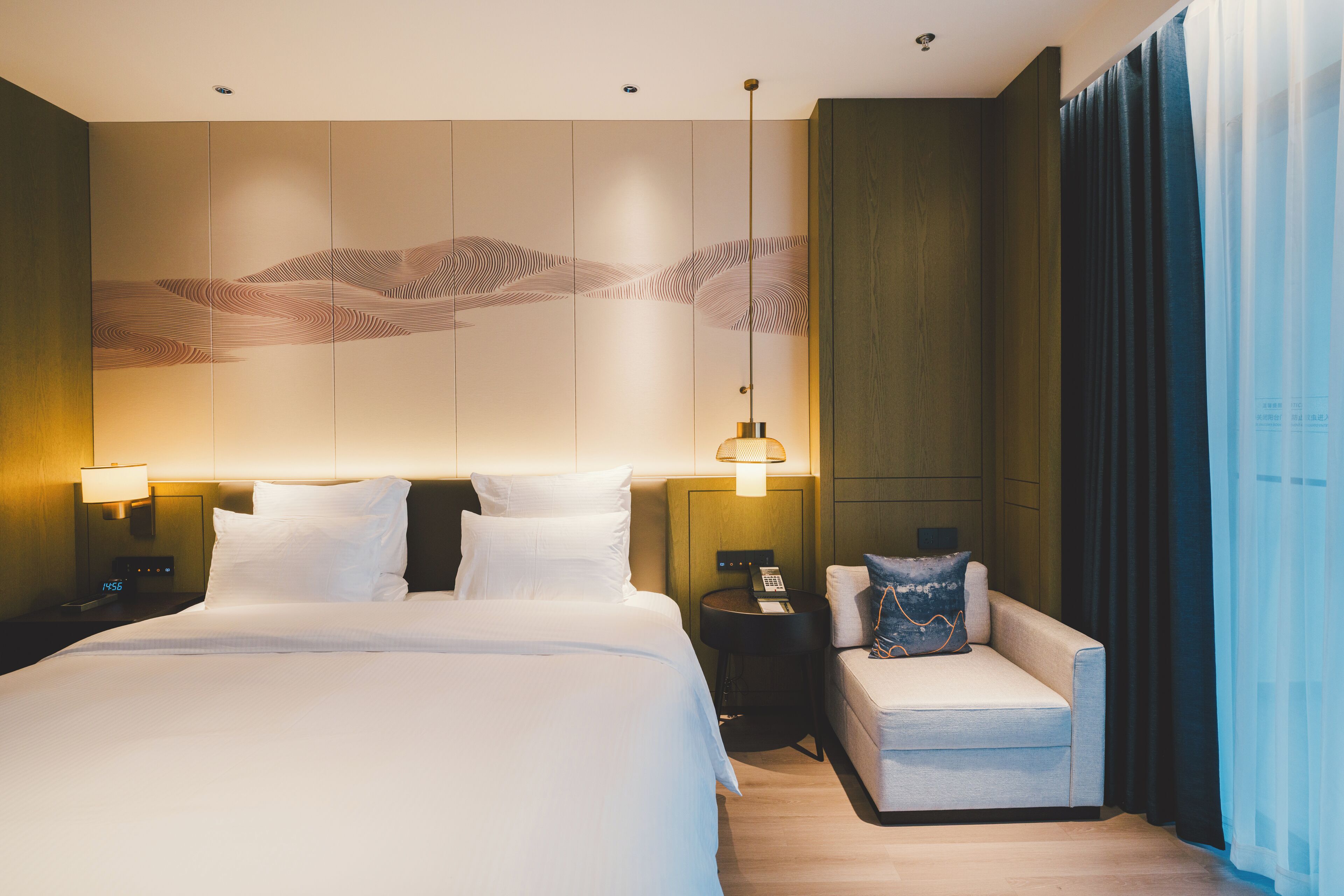 A hotel room with a large bed, a sofa, a nightstand with a lamp, and a wall with a decorative panel.