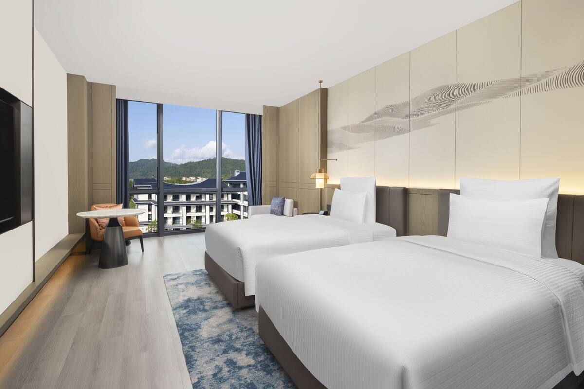 Ein modernes Hotelzimmer mit zwei Betten, einem großen Fenster mit Blick auf Gebäude, einem kleinen Tisch und einem Stuhl.