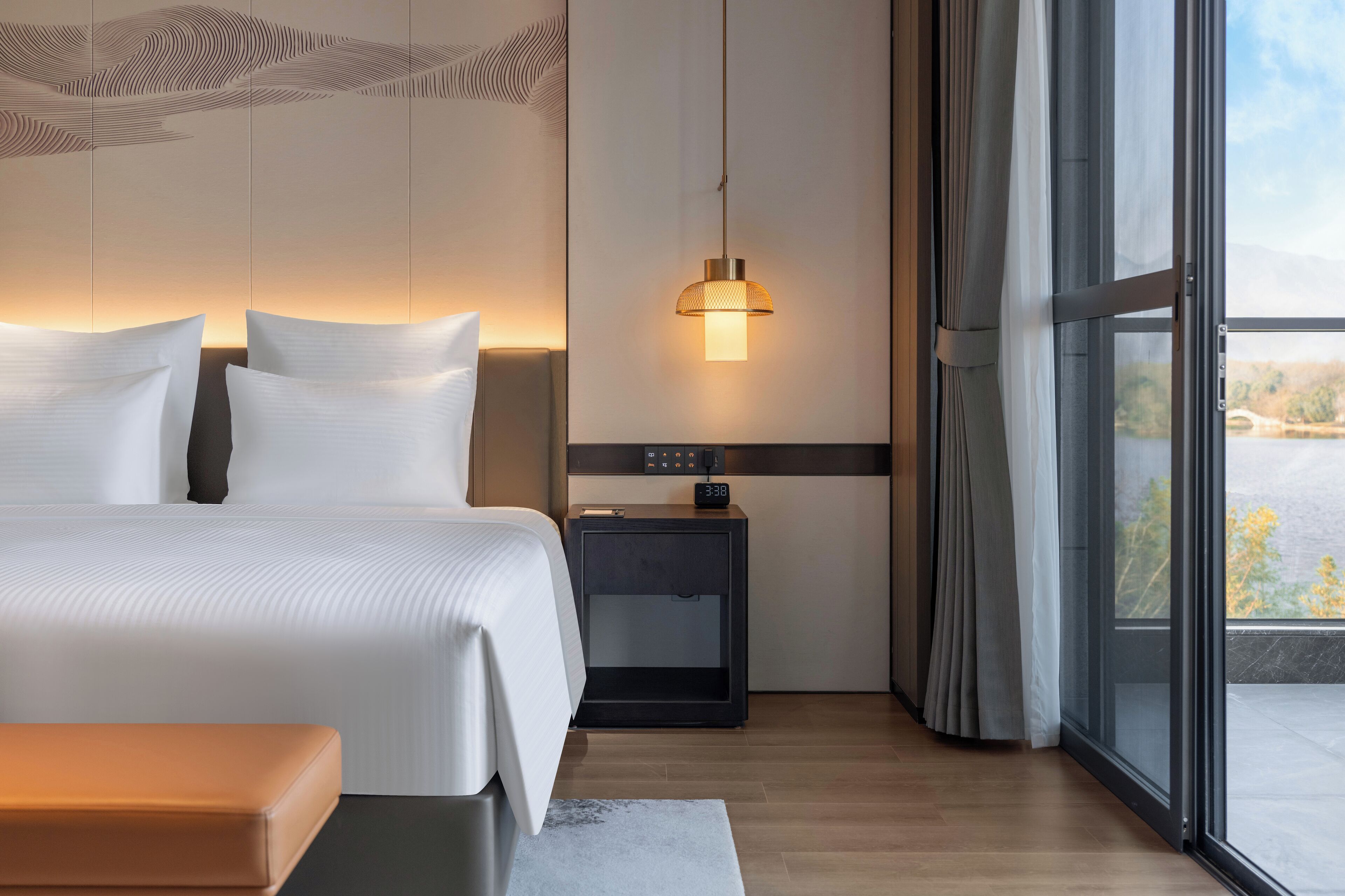 A modern hotel room with a large bed, a bedside table, a nightstand, and a view of a bridge and trees through a large window.