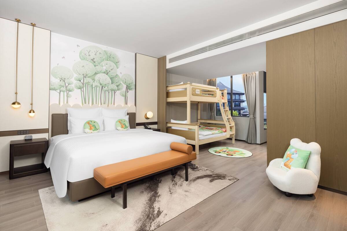 Ein modernes Hotelzimmer mit einem großen Bett, einem Schlafstockbett, einem weißen Sessel und einer Wandmalerei von Bäumen.