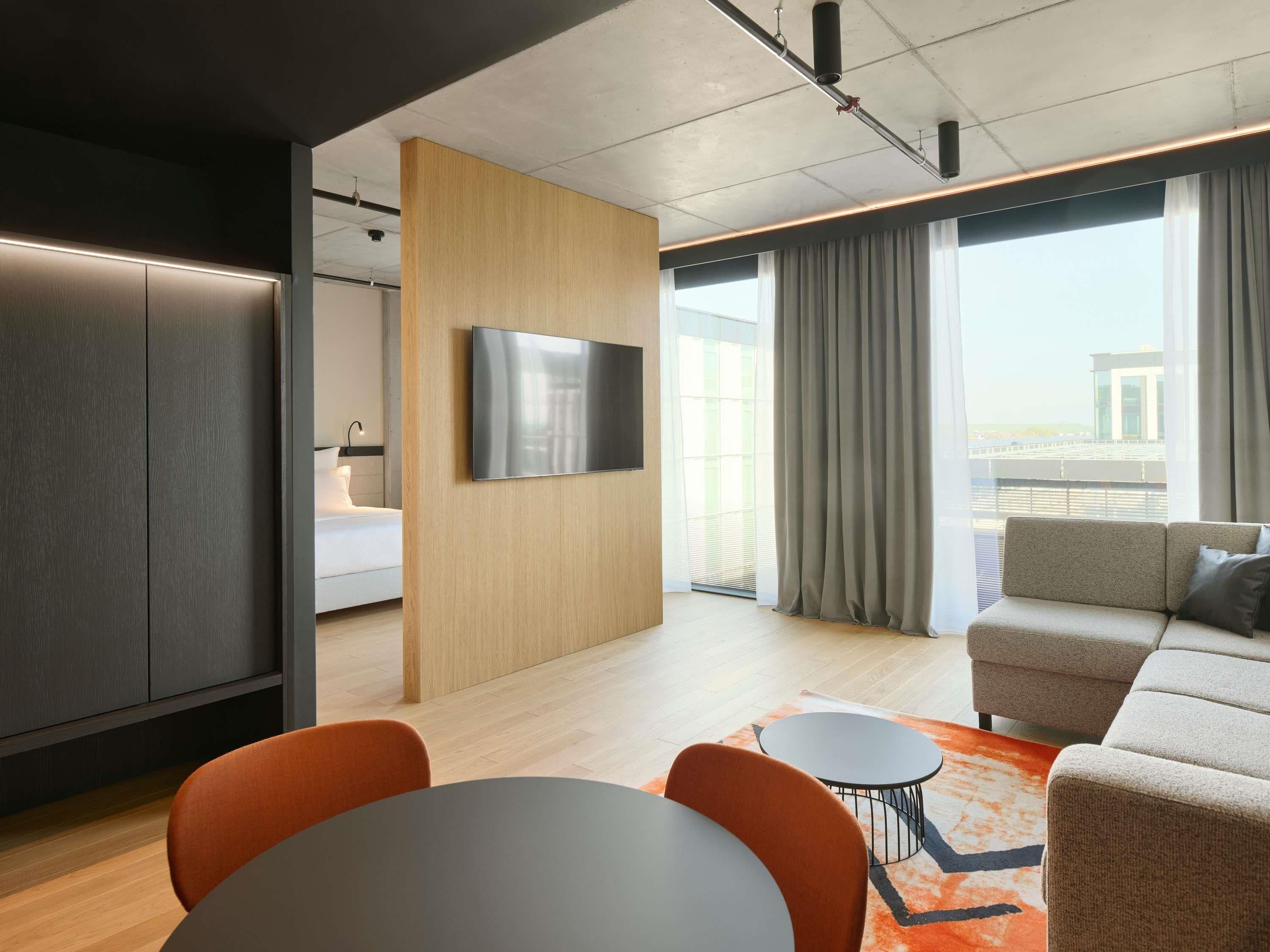 A modern hotel room with a flat-screen TV mounted on a wooden panel, a sofa, a dining table with chairs, and a large window with curtains.