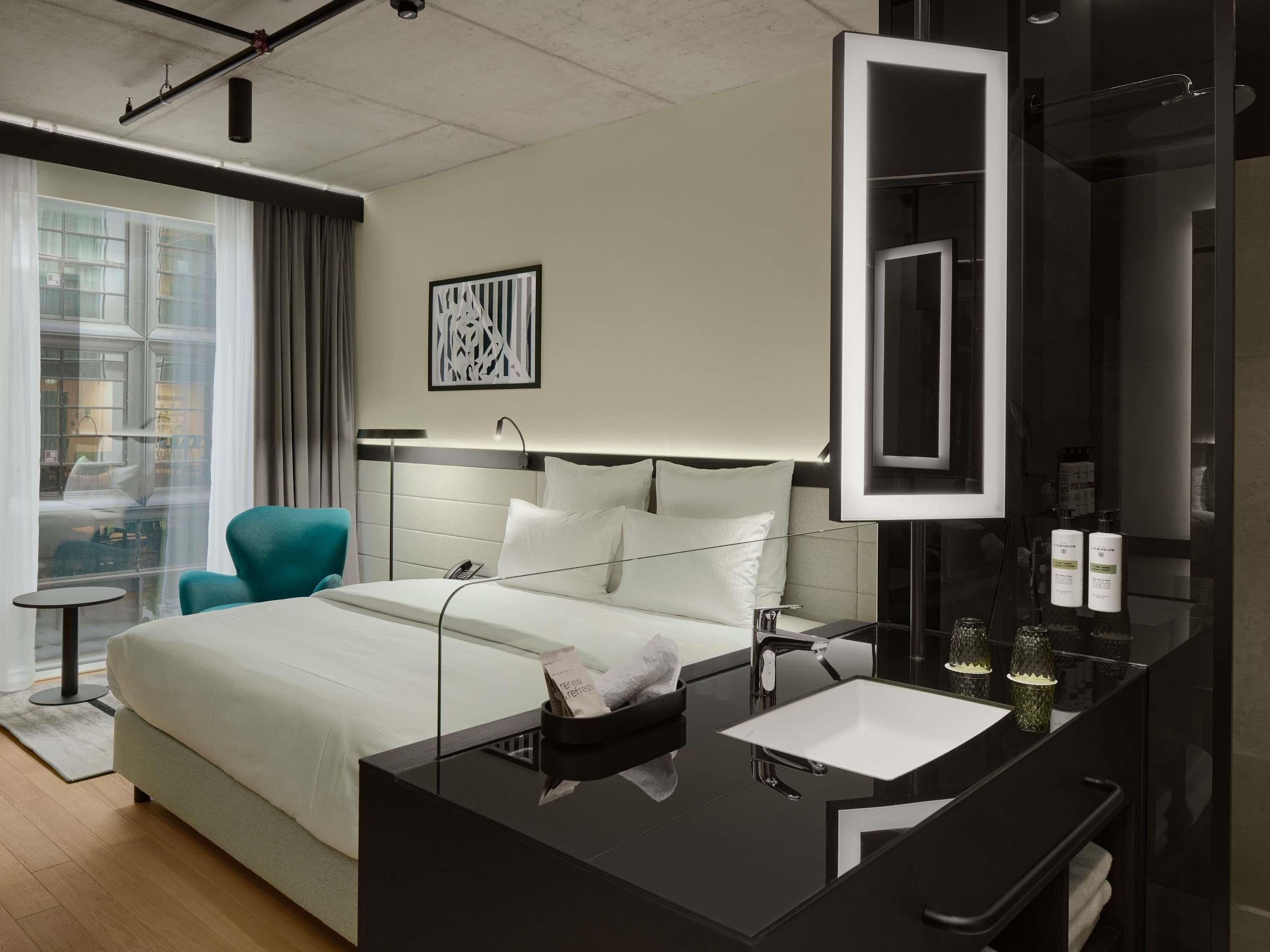 Una habitación de hotel moderna con una cama grande, una mesita de noche, un espejo y una ventana con cortinas.