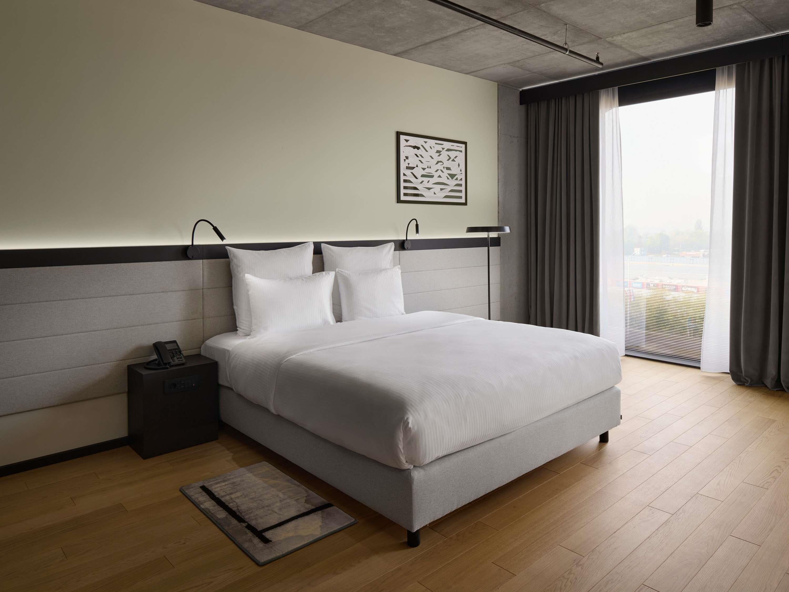 Una habitación de hotel moderna con una cama grande, una mesita de noche con teléfono, una alfombra pequeña y una ventana con cortinas.