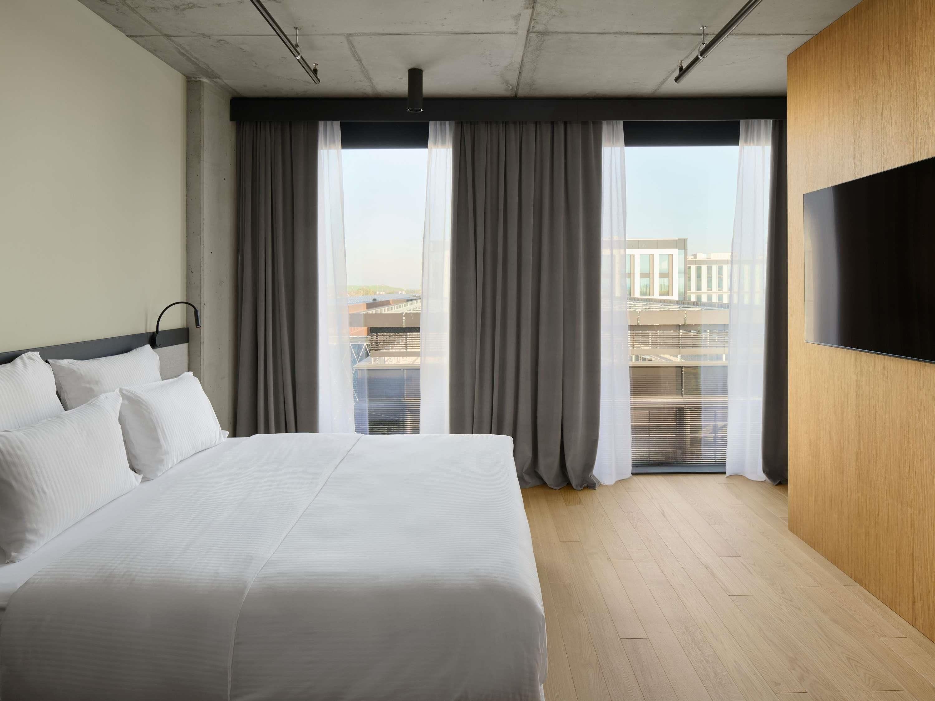 A modern hotel room with a large bed, a flat-screen TV mounted on the wall, and a view of the city through large windows.