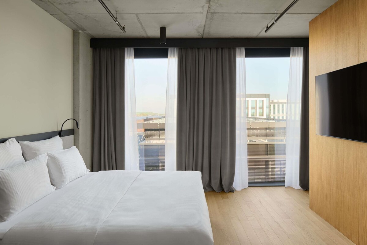 A modern hotel room with a large bed, a flat-screen TV mounted on the wall, and a view of the city through large windows.