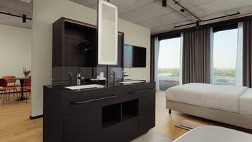 A modern hotel room with a large bed, a dark wooden vanity with a mirror, a flat-screen TV, and a city view through large windows.