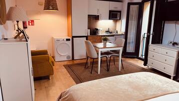 Een kleine hotelkamer met een bed, een eettafeltje, een wasmachine, een magnetron en een dressoir.