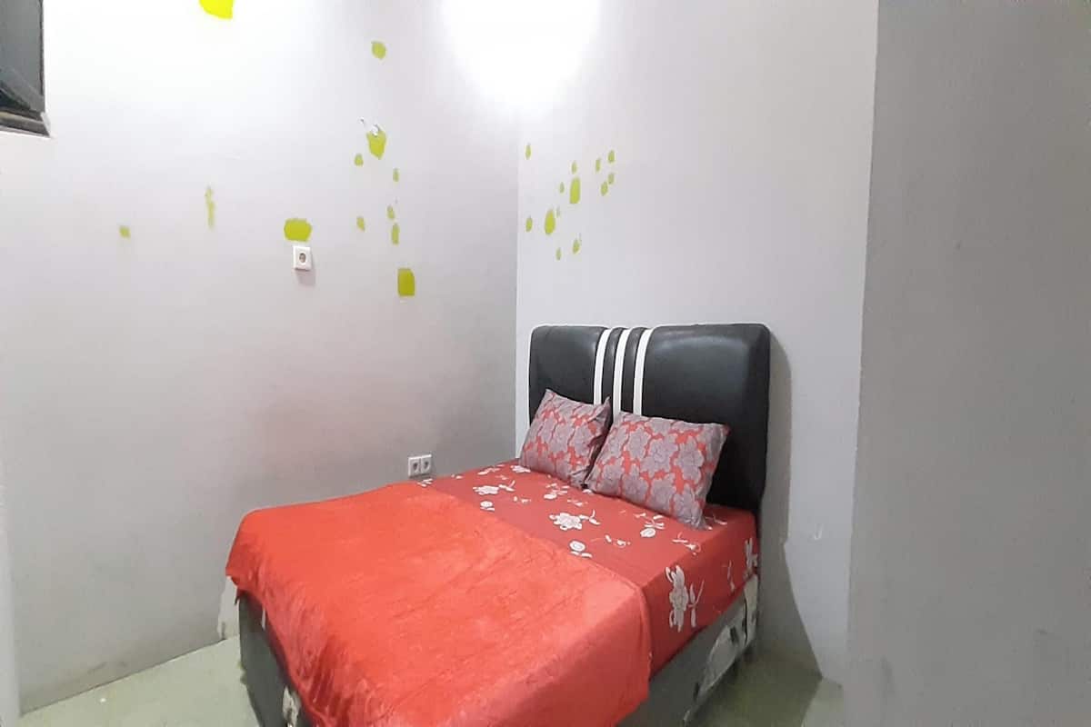 A small, neatly arranged bedroom with a single bed, a red bedspread, and a headboard with a white stripe.
