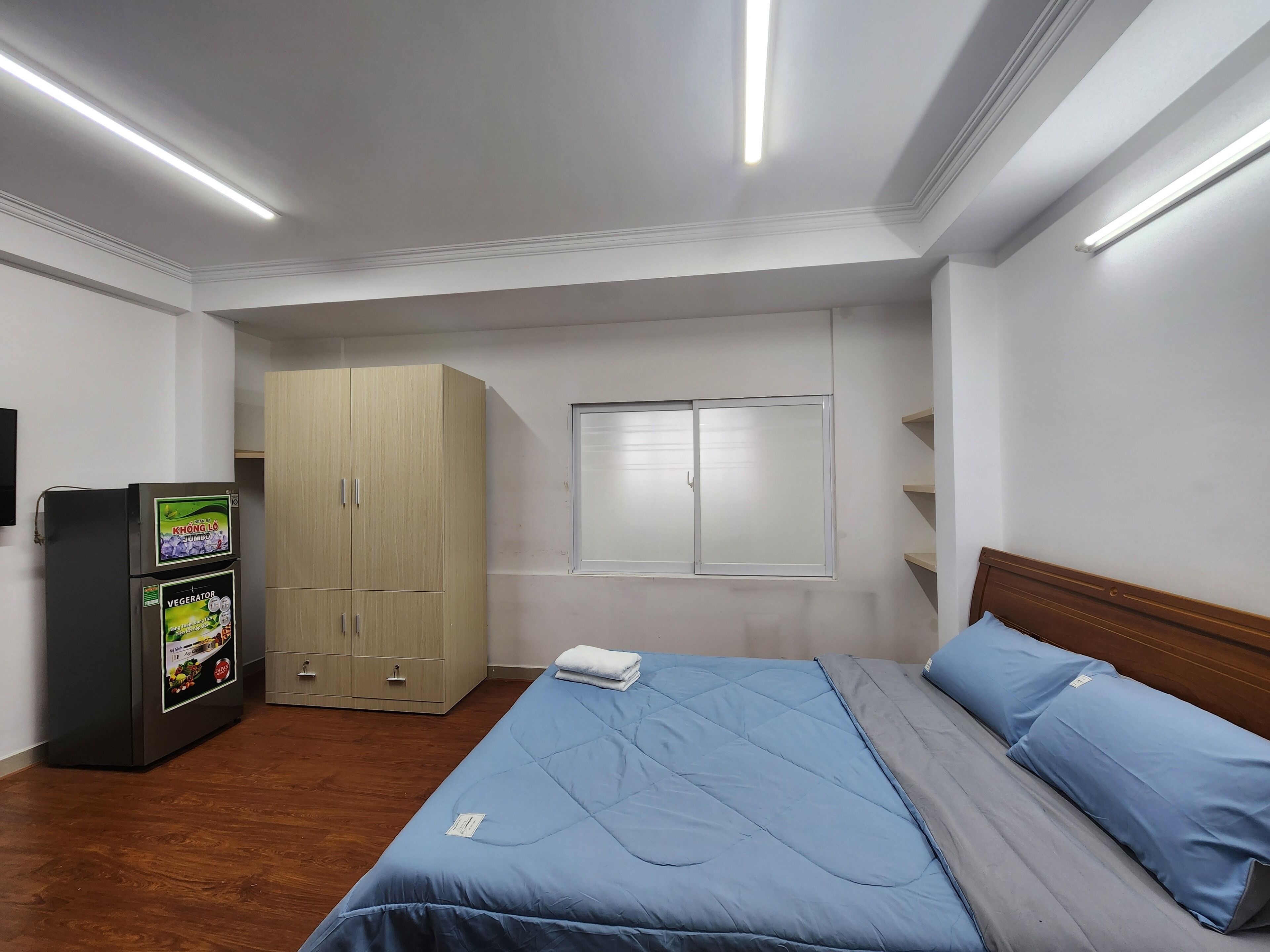 A bedroom with a bed, a wardrobe, a small refrigerator, and a window with blinds.