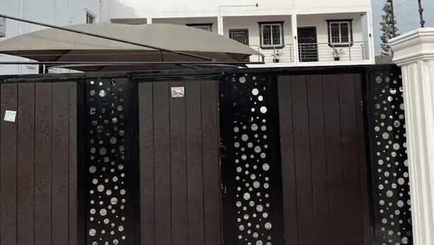 Ein von einem Metallzaun mit kreisförmigen Ausschnitten gesperrter Eingang, der zu einem weißen Gebäude mit Balkon führt.