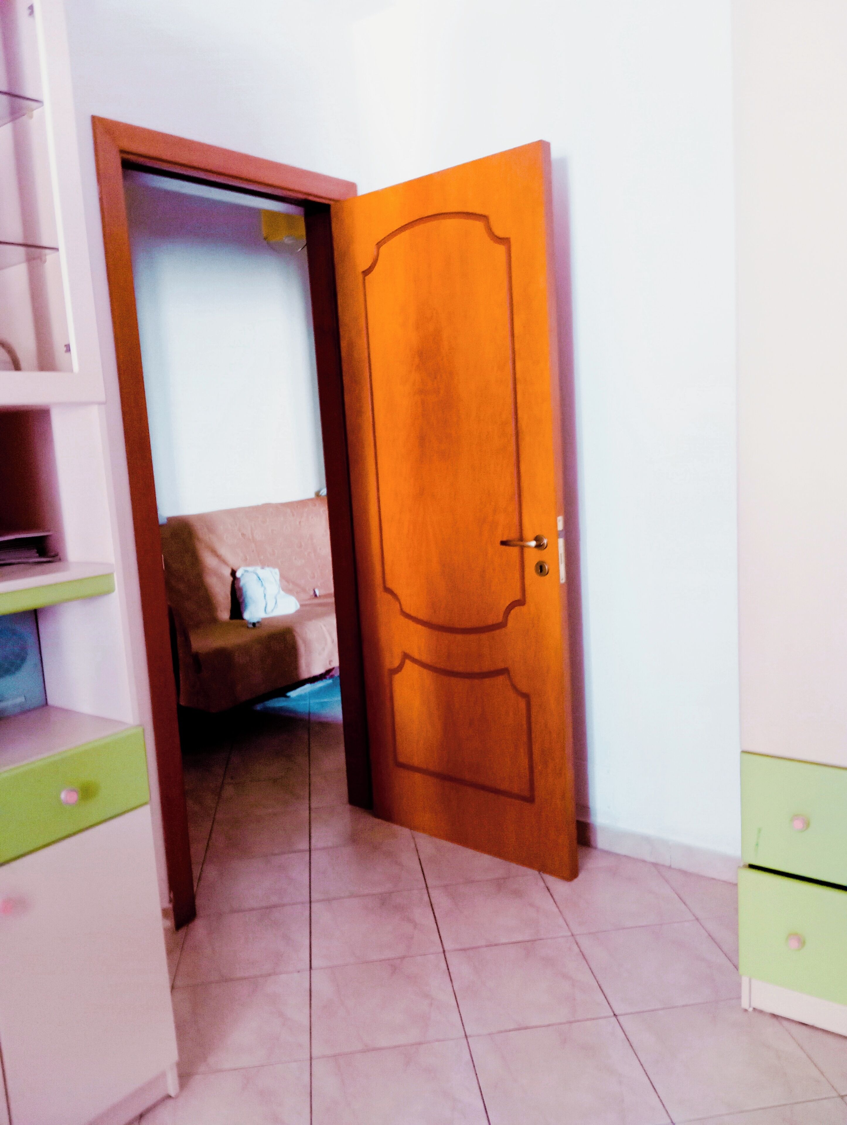 Une pièce avec une porte en bois, un lit, une commode verte et blanche, et un sol carrelé.
