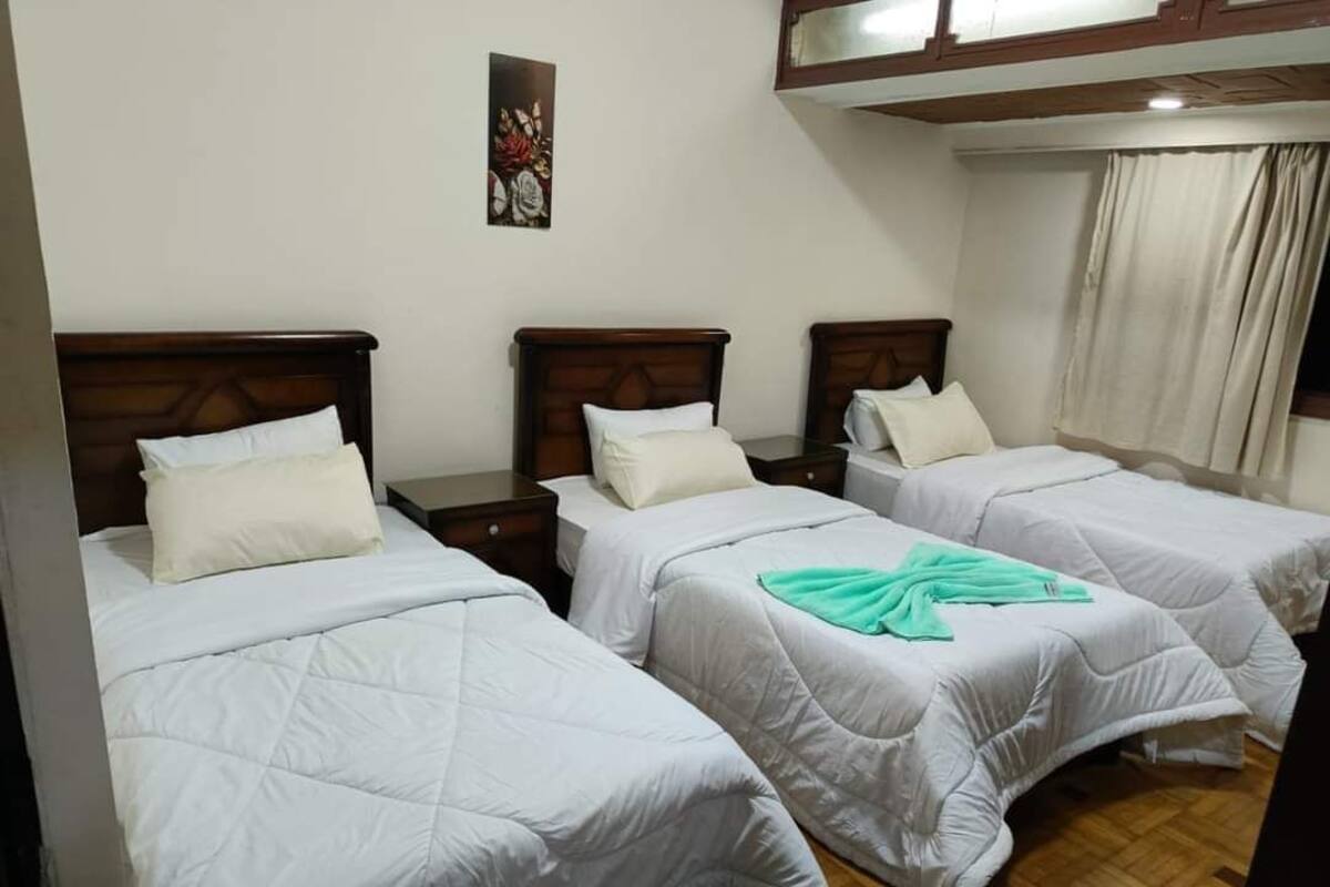 A hotel room with three single beds, each with a wooden headboard and white bedding. There is a painting on the wall above the beds and a window with curtains.