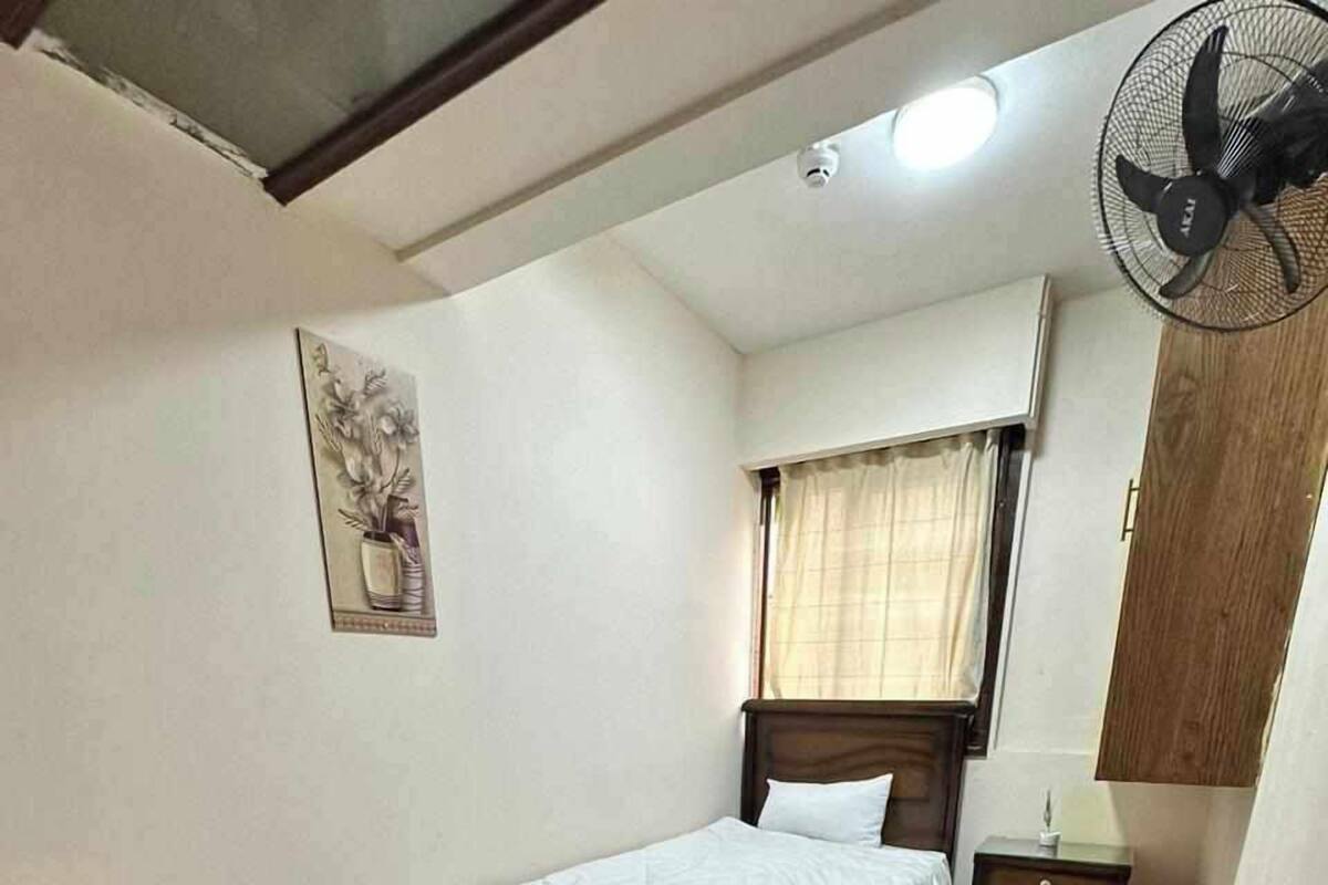 A small, neatly arranged bedroom with a single bed, a chair, a small table, and a wall-mounted fan.