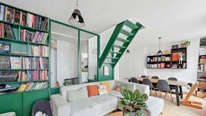 Un salon moderne avec un canapé gris, une bibliothèque verte, une table basse en bois et un escalier.