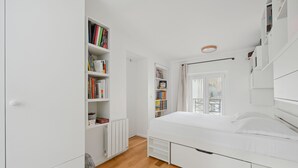 Une chambre avec un lit blanc, un plancher en bois, un radiateur, une bibliothèque et une fenêtre donnant sur un beau paysage.