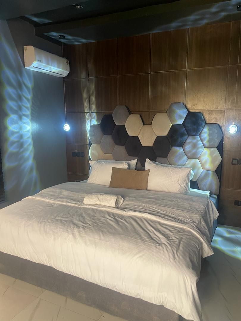 A modern bedroom with a hexagonal headboard, a bed with white linens, and an air conditioning unit mounted on the wall.