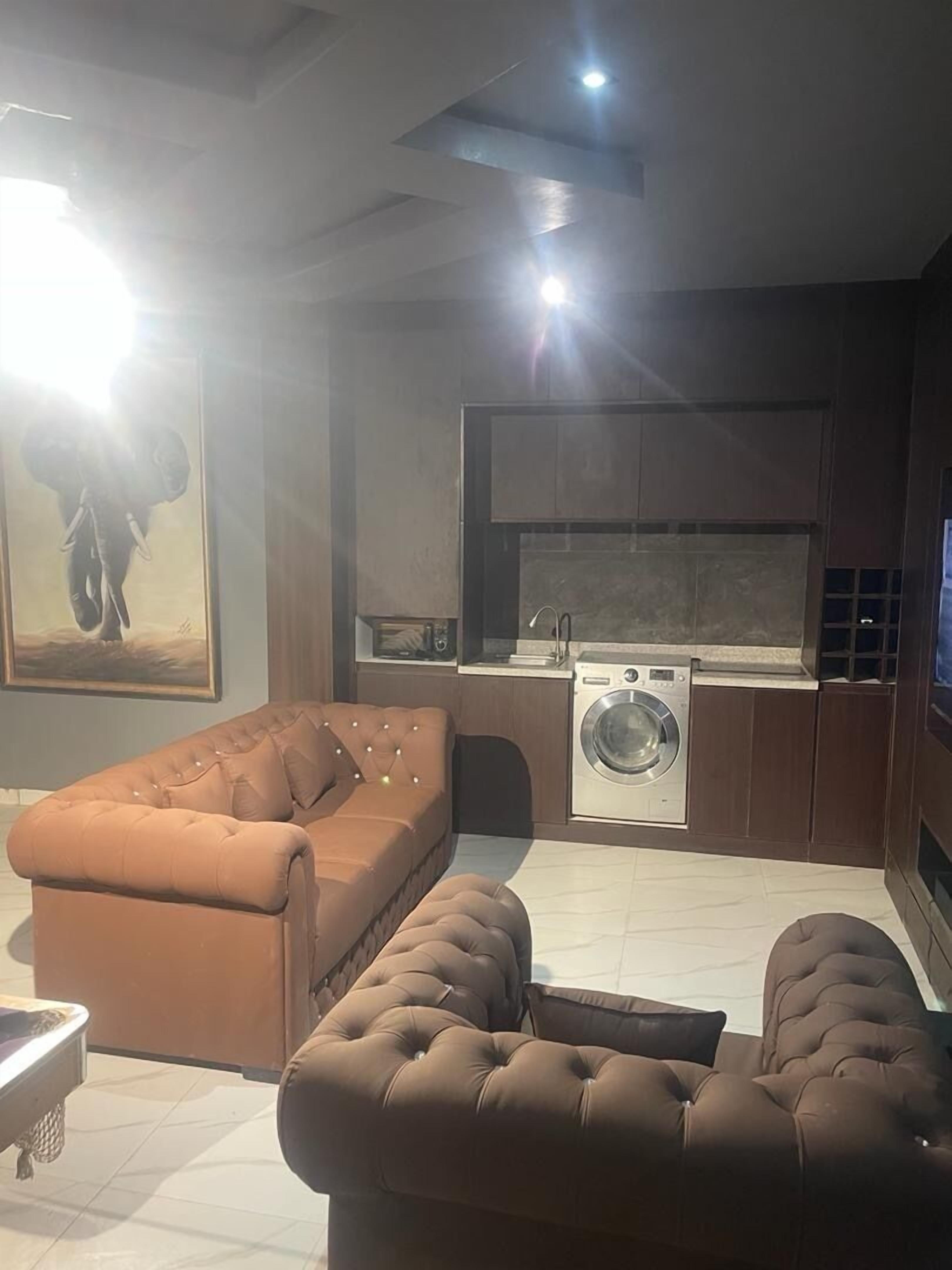 A modern living room with a large brown sofa and ottoman, a washing machine, and a kitchen area.
