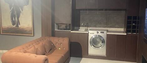 A modern living room with a large brown sofa and ottoman, a washing machine, and a kitchen area.