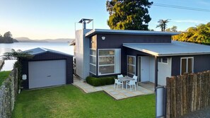 A modern house with a garage, a patio with chairs, and a view of the water. - Lakefront Oasis - Jetty, Hot Tub & Kayaks (Okere Falls)