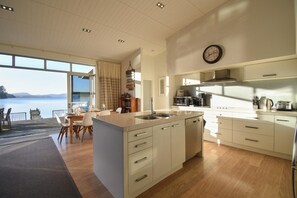 A modern kitchen with a large island, a dining area with a view, and a kitchen counter with appliances. - Lakefront Oasis - Jetty, Hot Tub & Kayaks (Okere Falls)