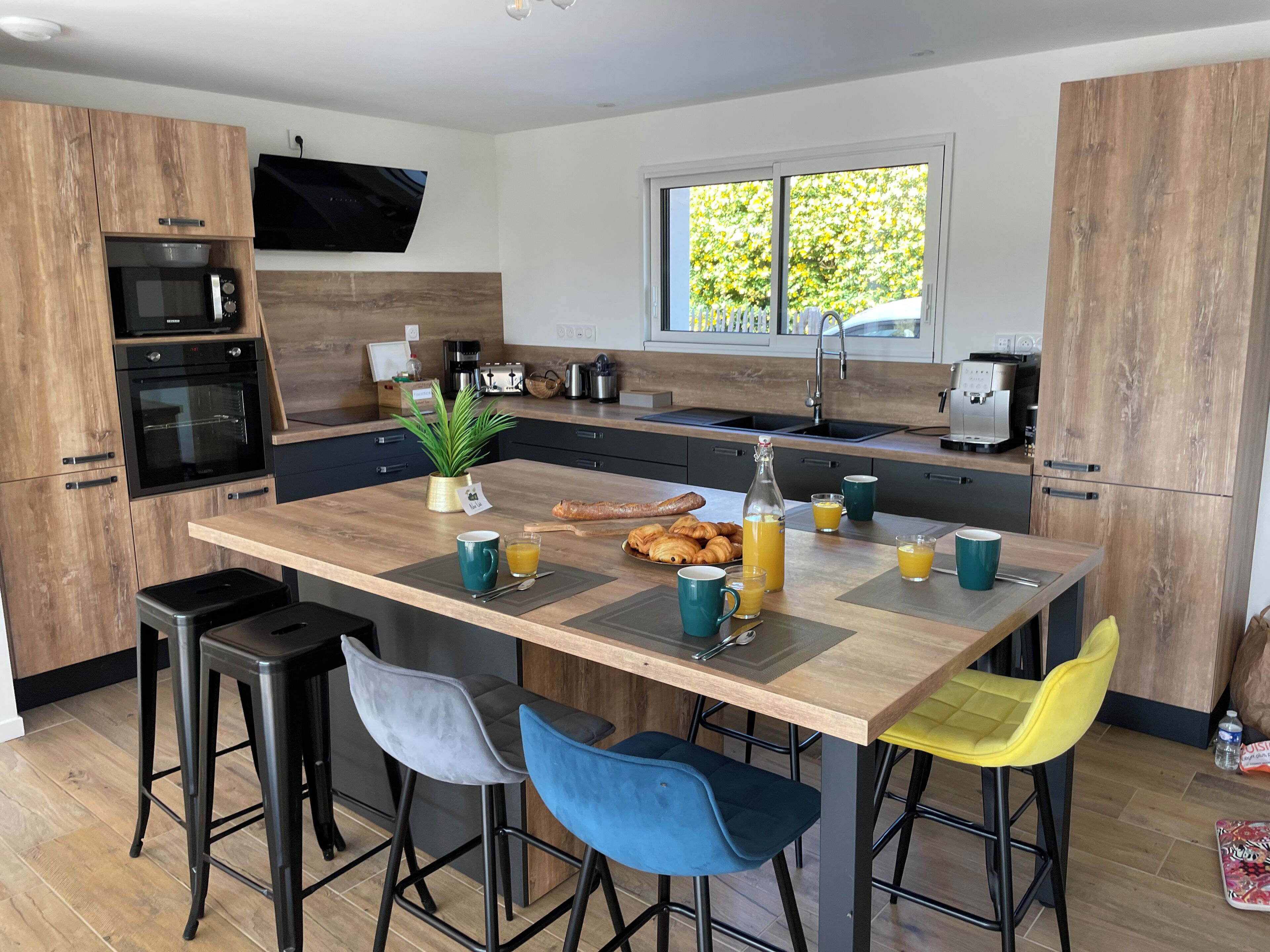 Een moderne keuken met houten kastjes, een centraal eiland met barkrukken, een televisie boven het eiland en een raam dat natuurlijk licht toelaat.