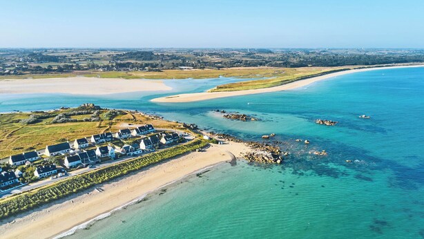 An aerial view of a coastal area with a sandy beach, clear blue waters, and a cluster of houses with distinct roofs.